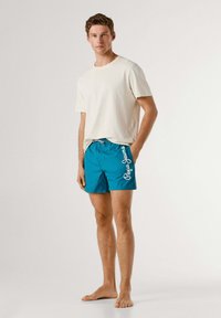 Ung mand stående barfodet iført en cremefarvet t-shirt og blå badeshorts med hvidt "Pepe Jeans"-logo på siden.