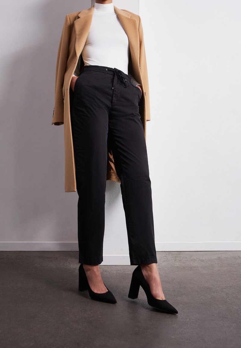 Femme portant un pantalon taille haute noir, des chaussures à talons hauts noires, un col roulé blanc, et un manteau couleur camel posé sur les épaules, debout à l'intérieur.