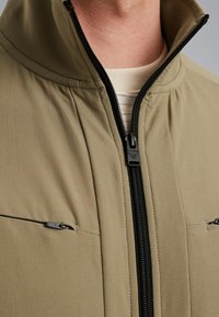 Giacca verde oliva con colletto alto, chiusura frontale con zip e due tasche sul petto con zip. Presenta una texture liscia e dettagli in zip nera.