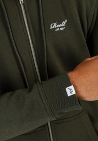 Sudadera con cremallera de color verde oscuro, hecha de una tela suave, con un logo blanco en el pecho y puños acanalados. Etiqueta notable en la muñeca.