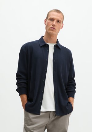 Mann trägt ein dunkelblaues Hemd mit Knöpfen über einem weißen T-Shirt und beigen Hosen, steht mit den Händen in den Taschen vor weißem Hintergrund.