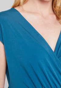 Türkises Kleid mit Wickeldesign, weicher Textur und kurzen Ärmeln. Es verfügt über einen tiefen V-Ausschnitt und eine geraffte Taille, die eine schmeichelhafte Silhouette andeutet.