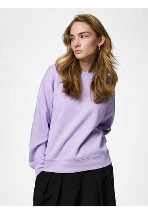 Jauna sieviete ar gariem viļņainiem matiem, valkājot sudraba riņķa auskarus, lavandas krāsas sporta kreklu un melnas bikses, skatās pa kreisi pret vienkrāsainu fonu.