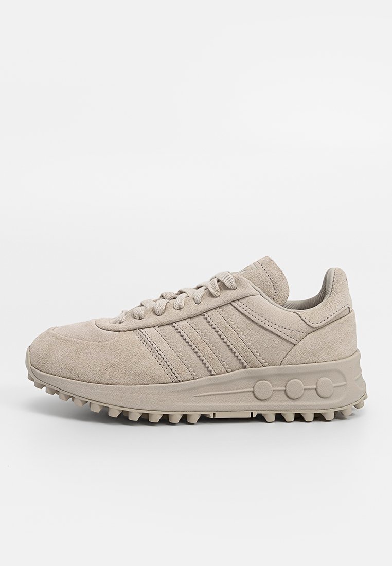 Beige suède sneaker met drie zijstrepen, ronde neus, gemêleerde bovenkant en een chunky rubberen zool met cirkelvormige accenten.