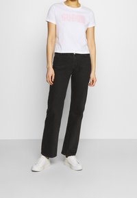 Hvid cropped t-shirt med "GUESS" logo i lyserød, parret med højtaljede sorte jeans og hvide sneakers. Enkel, afslappet outfit.
