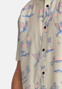 Camisa de botones de manga corta en beige claro con estampado tropical en azul y rosa, que presenta palmeras y pájaros. Incluye un bolsillo en el pecho y botones negros.