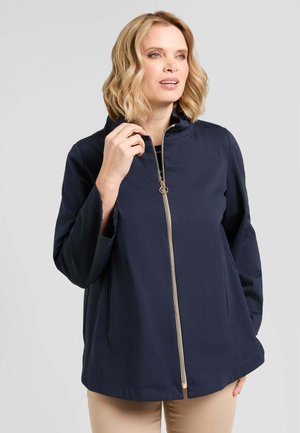 Donna con capelli biondi che indossa una giacca blu navy con zip e collo alto e pantaloni beige, mentre tiene la cerniera vicino al collo.