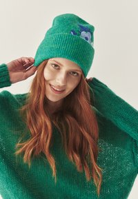 Căciulă verde tricotată cu textură rib și broderie florală. Modelul poartă un pulover verde asortat, cu croială lejeră și țesătură texturată.