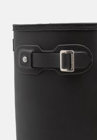 Bota de goma negra con acabado mate, que presenta un detalle de hebilla de metal plateado y un borde superior texturizado. Diseño durable e impermeable.