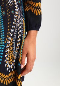 En kvinnas hand vilar bredvid en mönstrad svart klänning dekorerad med blå, vita och gyllene blad- och vinmönster.