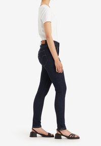 Vaqueros ajustados azul oscuro hechos de mezclilla, con cintura alta, bolsillos traseros y una etiqueta de cuero con la marca en la cintura. Combinados con una camisa blanca y sandalias negras.