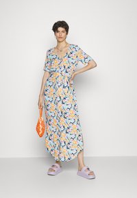 Vestido maxi floral com mangas curtas, apresentando um fundo azul-marinho e flores brilhantes em laranja, azul e rosa. Combinado com sandálias lilás e uma bolsa laranja.