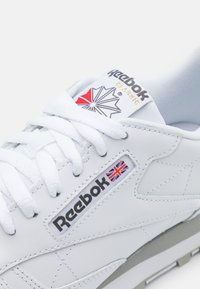 Weiße Reebok-Sneaker mit Obermaterial aus Leder, ausgestattet mit Schnürverschluss, Logotags, dreieckigen Akzenten und grauer Gummisohle.