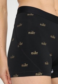 Shorts de sport noirs en tissu extensible, ornés d'un motif répétitif du logo "nike" en or. Ils ont une coupe ajustée et des bords sans couture.