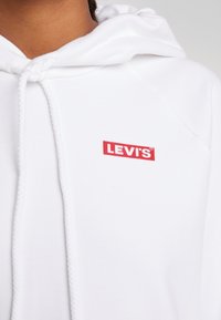 Sweat à capuche blanc avec un logo rouge "LEVI'S". Il possède une capuche avec cordon, une texture douce et des détails côtelés près de l'encolure.