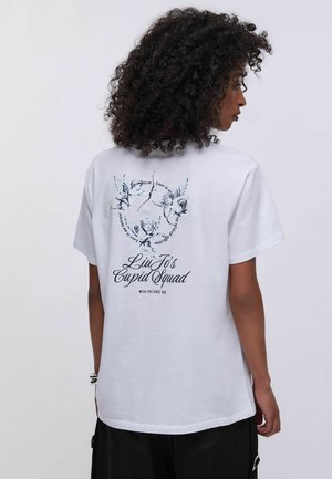 Femme aux cheveux bouclés portant un T-shirt blanc affichant trois chérubins avec des arcs et le texte "Liu Jo's Cupid Squad" dans le dos.