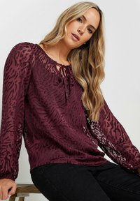 Blusa bordeaux con stampa animale testurizzata, maniche lunghe trasparenti, polsini elastici e dettaglio con nodo sul colletto. Tessuto morbido.