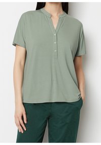 Mujer vestida con una blusa de botones de manga corta color verde claro con cuello mao y pantalones verde oscuro, con la mano en el bolsillo.