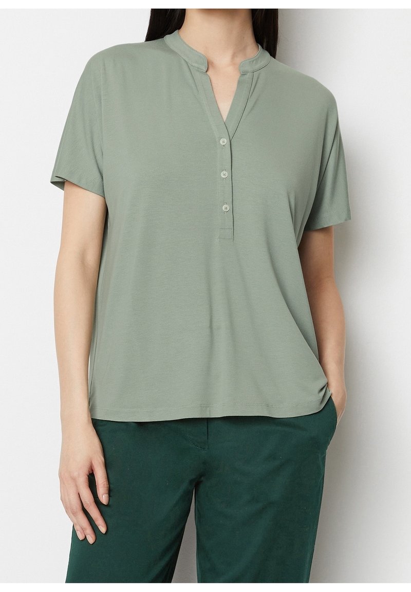 Mujer vestida con una blusa de botones de manga corta color verde claro con cuello mao y pantalones verde oscuro, con la mano en el bolsillo.