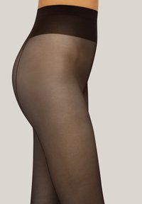 Torso e gambe di una donna che indossa collant neri trasparenti con un ampio elastico opaco su uno sfondo semplice.