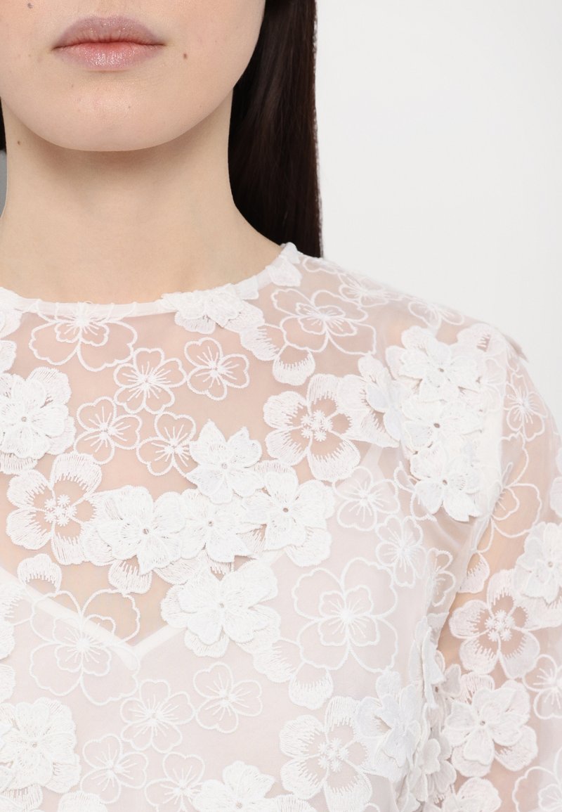 Witte bloemen-geëmbroide top van doorschijnende stof, met driedimensionale bloemen en een ronde halslijn. Gestructureerde en delicate ontwerpelementen.