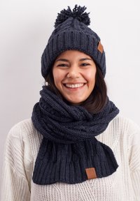 Navy knitted beanie with a pom-pom and matching scarf featuring ribbed texture and a brown label detail. Worn by a smiling person.