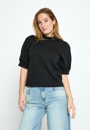 Femme aux longs cheveux blonds portant un haut noir à manches bouffantes et un jean bleu clair, debout devant un fond blanc uni.