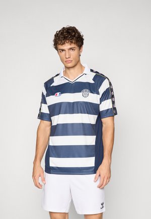 Man wearing navy and white striped sports jersey with logos and white shorts, standing against a plain light background.