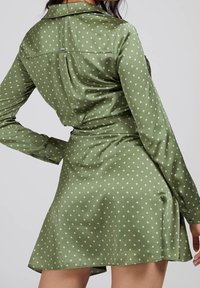 Grünes Kleid mit Polka-Dots, Kragen, langen Ärmeln und einem ausgestellten Rock. Glatte Textur; verfügt über ein dezentes Rückendetail und Knöpfe.