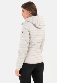 Lichtbeige pufferjack met capuchon, gequilt ontwerp en elastische manchetten. Bevat een klein bruin logo op de linkermouw.