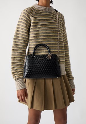 Personne portant un pull beige à rayures et une jupe plissée marron, portant un sac à main matelassé noir Guess avec une bandoulière en chaîne et des poignées supérieures.