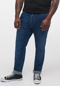 Jeans din denim de culoare albastru închis cu o croială dreaptă, având cusături deschise la culoare, un design clasic cu cinci buzunare și manșete răsucite. Asortați cu adidași negri cu glezna înaltă.