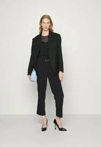 Mulher de pé, vestindo um blazer preto, uma blusa transparente com bolinhas, calças pretas justas e sapatos de salto pretos, segurando uma bolsa azul clara contra um fundo simples.