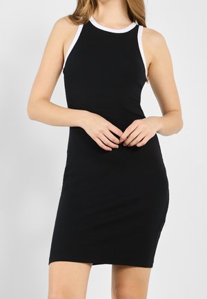 Femme portant une robe noire sans manches avec des bordures blanches, debout les mains légèrement jointes devant elle, sur un fond clair uni.