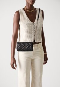 Borsa a tracolla in pelle nera trapuntata con hardware argentato, caratterizzata da un logo triangolare. Indossata con un gilet in maglia e jeans chiari.