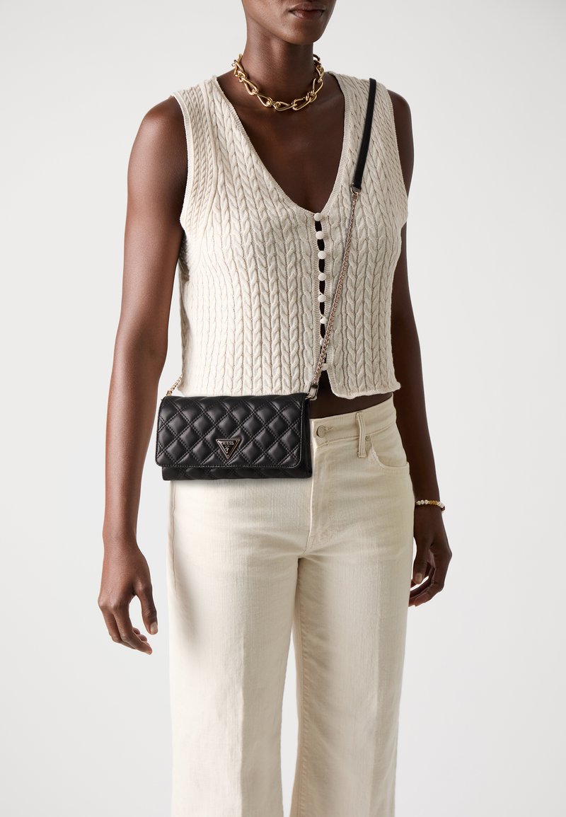 Borsa a tracolla in pelle nera trapuntata con hardware argentato, caratterizzata da un logo triangolare. Indossata con un gilet in maglia e jeans chiari.