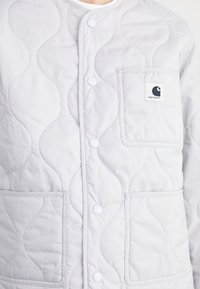 Jasnoszara pikowana kurtka z okrągłym dekoltem, zatrzaskami i kieszenią na piersi z naszywką z logo. Gładka faktura i widoczne przeszycia.