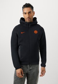 Sudadera con capucha negra, con cremallera, hecha de tejido suave. Presenta un logo de Nike naranja y un emblema redondo naranja, con bolsillos laterales y puños ajustados.