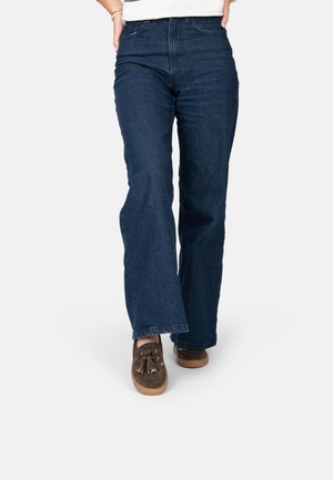 Jeans in denim blu scuro a zampa d'elefante con vita alta, caratterizzati da un taglio dritto e orlo pulito. Abbinati a scarpe stile mocassino marroni.