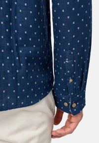Camisa de color azul marino con un sutil patrón de cuadrados blancos, hecha de tejido ligero, con puños abotonados y un dobladillo curvado.