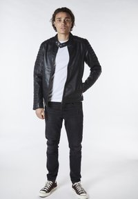 Chaqueta de cuero negra con hombros acanalados, camiseta blanca, jeans negros desgastados y zapatillas oscuras, de pie contra un fondo neutro.