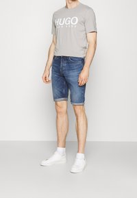 Miestny svetlošedý bavlnený tričko s logom "HUGO" v bielej farbe, v kombinácii s modrými džínsovými šortkami s ohrnutými rukávmi a bielymi teniskami.