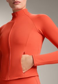 Oranžová dlhá fitness bunda s dlhým rukávom, vysokým golierom, predným zipsom a bočnými vreckami, vyrobená z hladkej, elastickej textílie.