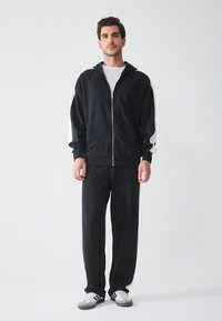 Zwarte rits-hoodie met witte zijstrepen, gecombineerd met zwarte joggingbroek. Het materiaal lijkt zacht en lichtjes getextureerd. Sneakers zijn grijs en wit.