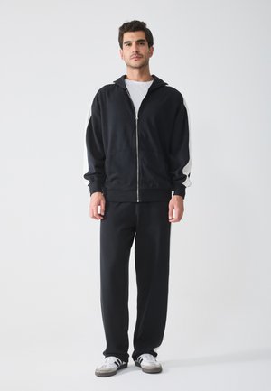 Felpa nera con zip e strisce laterali bianche, abbinata a pantaloni felpati neri. Il materiale sembra morbido e leggermente_TEXTURIZZATO. Le sneaker sono grigie e bianche.