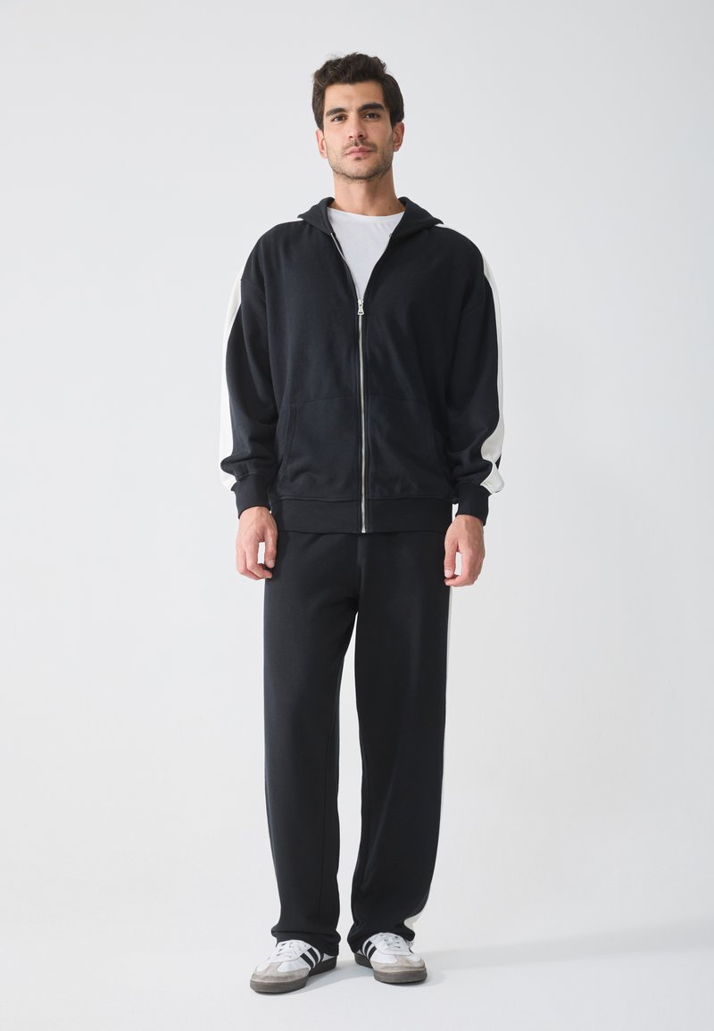Zwarte rits-hoodie met witte zijstrepen, gecombineerd met zwarte joggingbroek. Het materiaal lijkt zacht en lichtjes getextureerd. Sneakers zijn grijs en wit.