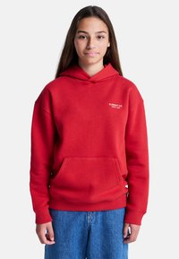 Giovane con capelli lunghi e castani, indossa una felpa rossa con la scritta "Element Co. Since 1992" e jeans blu, in piedi su uno sfondo bianco.