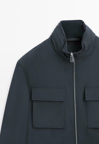 Veste zippée gris foncé avec col montant et deux grandes poches à rabat sur la poitrine, présentée sur un fond blanc.