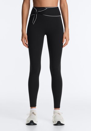 Kvinde iført sorte højtaljede leggings med bindebælte, hvide sokker og hvide sportssko, stående mod en ensfarvet baggrund.