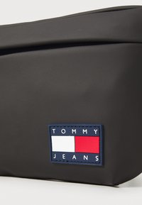 Fekete, matt anyagú tasak, amelyen egy téglalap alakú Tommy Jeans logó folt található kék, piros és fehér elemekkel, zárt kialakítású, látható fémalkatrészek nélkül.